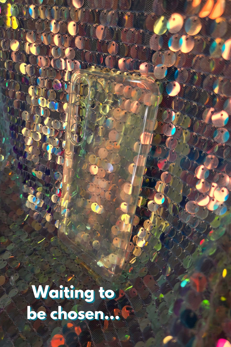 Close-up of a transparent phone case on a  glittery, sequined surface with text 'Waiting to be chosen...'.